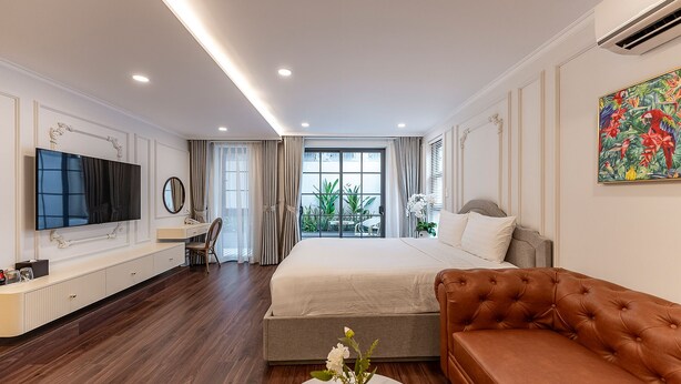 A modern hotel room with a large bed, a brown leather sofa, a TV, a desk, and a view of greenery through the window. - Golden Sun Apartment & Hotel (Ho Chi Minh City)