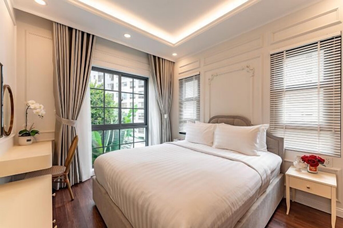 Een slaapkamer met een groot bed, een nachtkastje, een stoel, een raam met gordijnen en een kamerplant.