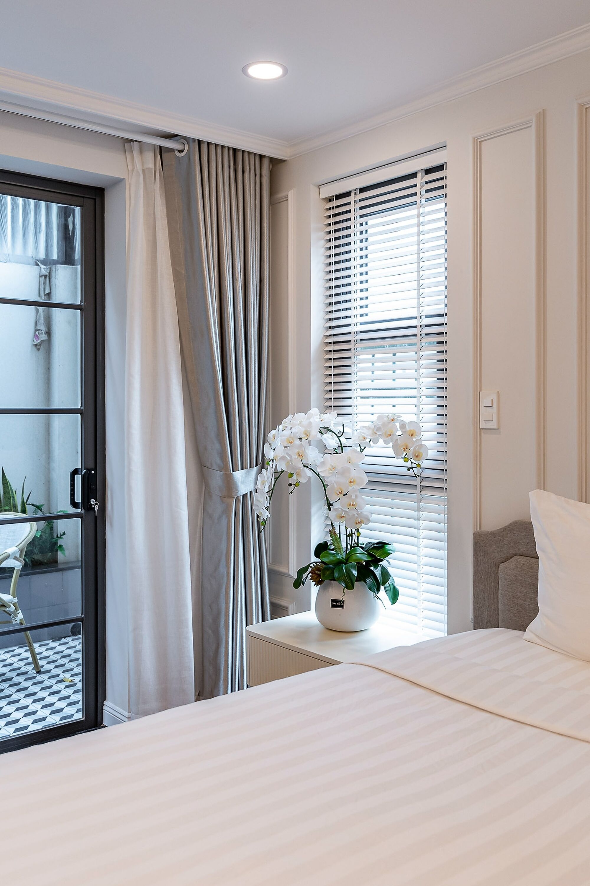 A hotel room with a bed, a window with blinds, a potted plant with white flowers, and a view of the outside balcony.