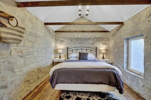 A bedroom with a stone wall, a wooden ceiling, a bed with a headboard, bedside tables with lamps, and a window.