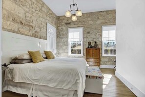 A bedroom with a stone wall, a bed with white bedding, a wooden nightstand, and a chandelier. - The Homestead Estate (Fredericksburg)