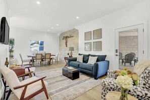A modern living room with a blue sofa, a wooden coffee table, and a patterned armchair. There is a dining area with a table and chairs, and a stone accent wall. - The Homestead Estate (Fredericksburg)