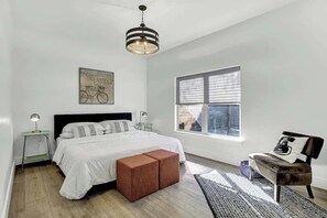 A bedroom with a bed, a nightstand, a bench, a window, and a hanging light fixture.