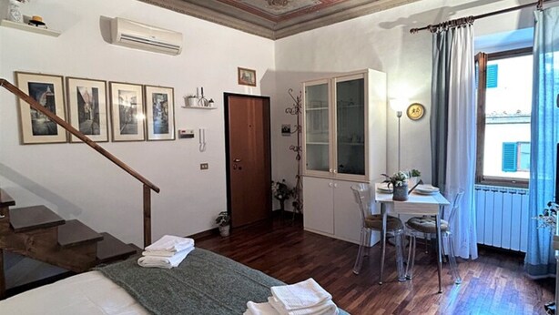 A room with a wooden floor, a bed with a green blanket, a desk with a chair, a window with curtains, and a decorative ceiling. - Romantic Nest Florence Studio (Firenze)