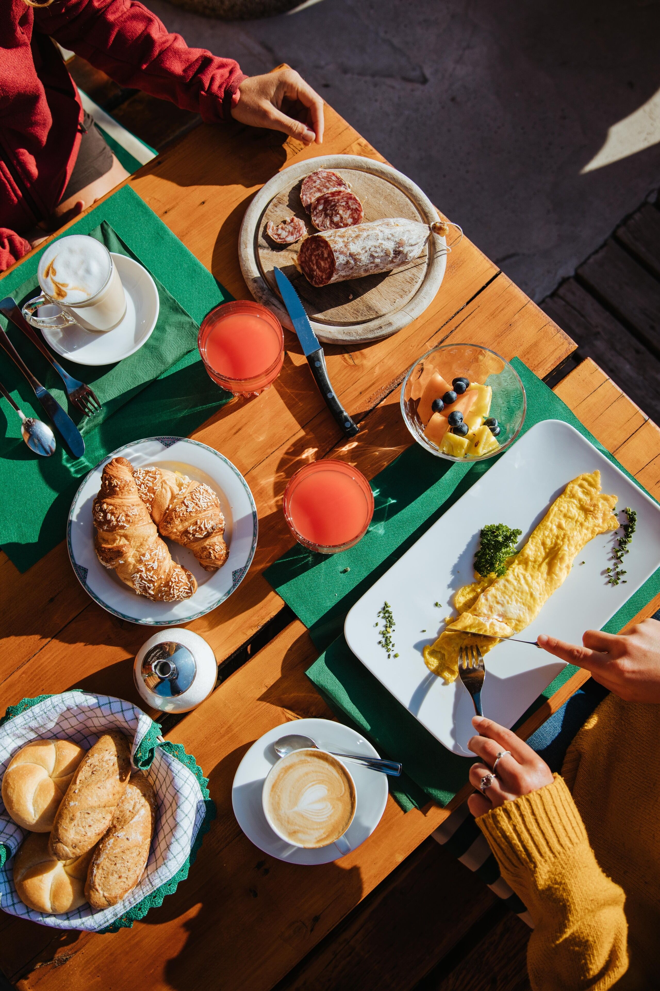 A wooden table with breakfast items including croissants, a plate of sliced salami, a bowl of fruit, and a plate of omelette.
