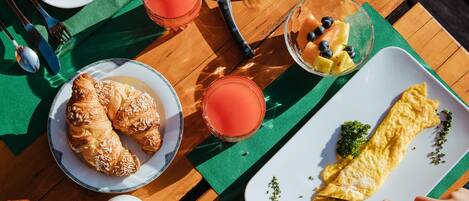 A wooden table with breakfast items including croissants, a plate of sliced salami, a bowl of fruit, and a plate of omelette.