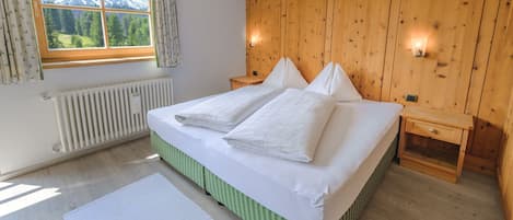 Una camera da letto con una testiera in legno, un'ampia finestra vista montagna, un termosifone e un comodino.