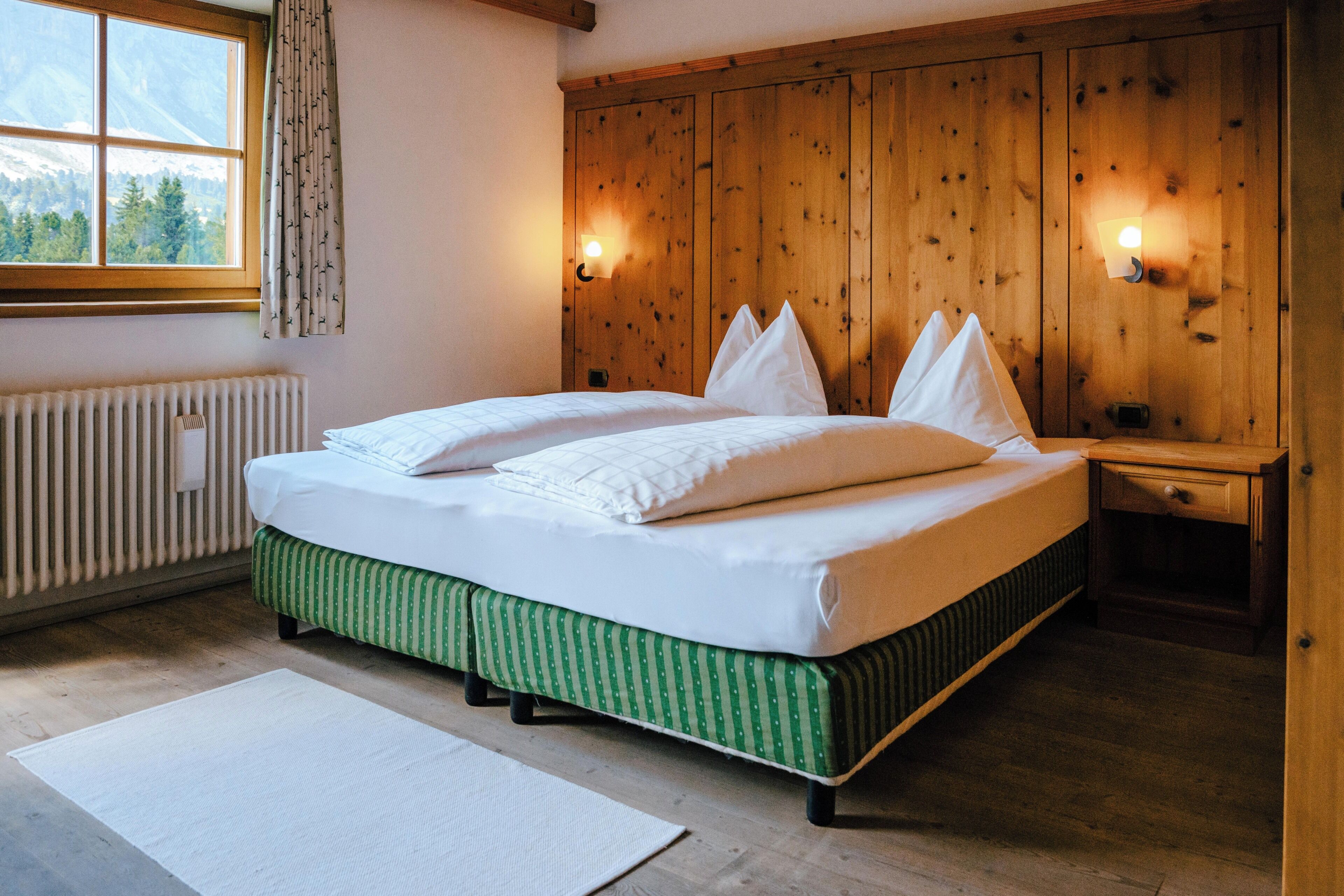 Un dormitorio con cabecera de madera, cama con ropa blanca, un banco verde a rayas, un radiador blanco y una ventana con vista a árboles.