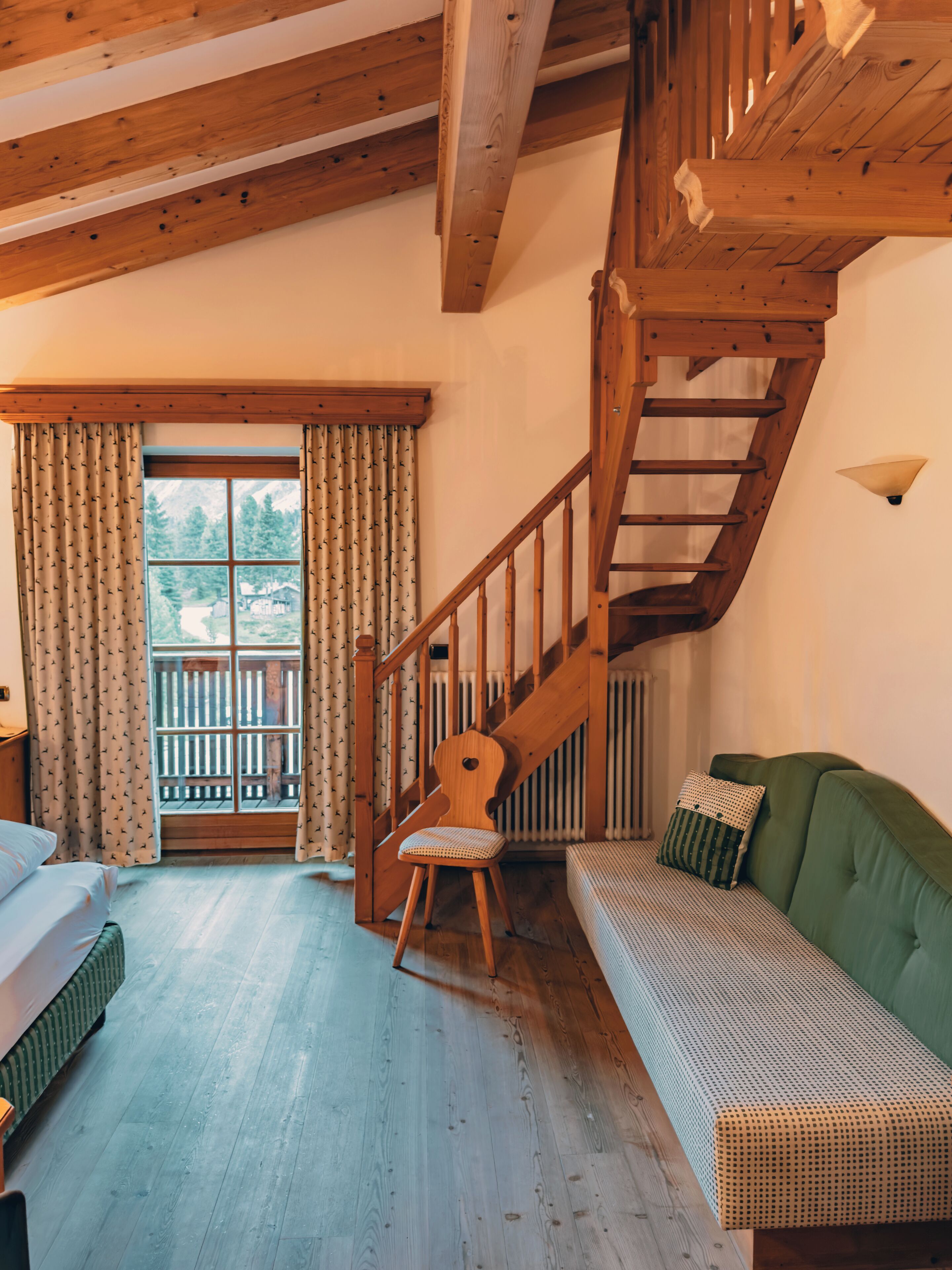 Una habitación con escalera de madera, un sofá verde, un sillón de balancín de madera y una ventana con cortinas.