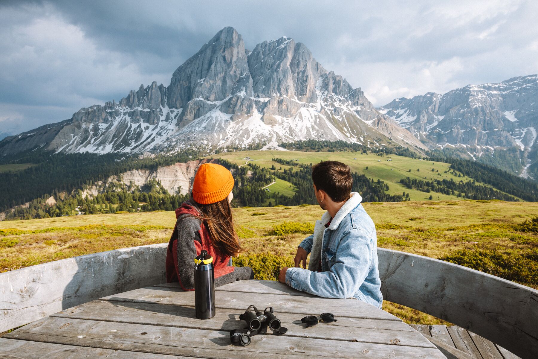 Two people sitting on a wooden bench with a mountain view in the background.