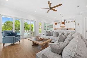A spacious living room with a large grey sofa, a blue armchair, and a wooden coffee table. There is a ceiling fan, a kitchen area with white cabinets, and large windows offering a view of the outdoors.