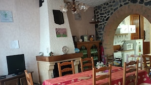 A dining area with a red tablecloth, wooden chairs, a stone wall, a kitchen visible through an archway, and a television on a cabinet. - Gîte Chez Mimi à Anor (Anor)