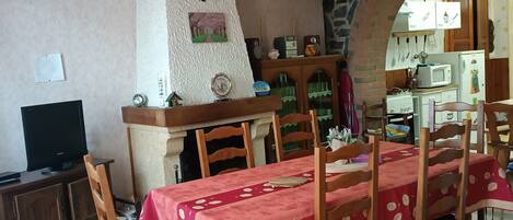 Un coin repas avec une nappe rouge, des chaises en bois, un mur en pierre, une cuisine visible à travers une arche et un téléviseur posé sur un meuble.