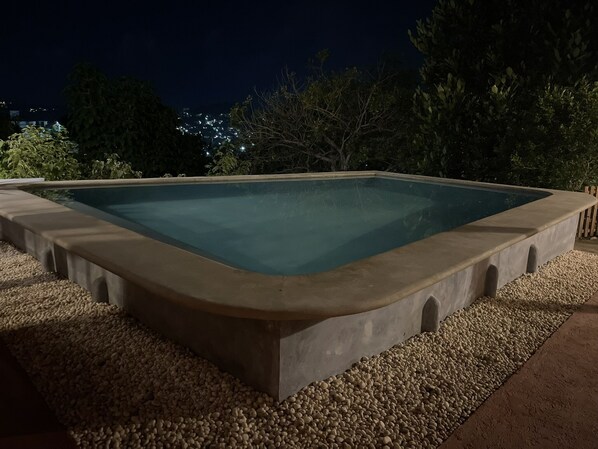 Una piscina rectangular al aire libre rodeada de un borde de piedras, con árboles y un cielo nocturno al fondo.