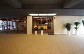 Entrada de hotel con puerta de cristal, plantas en macetas y suelo de baldosas.