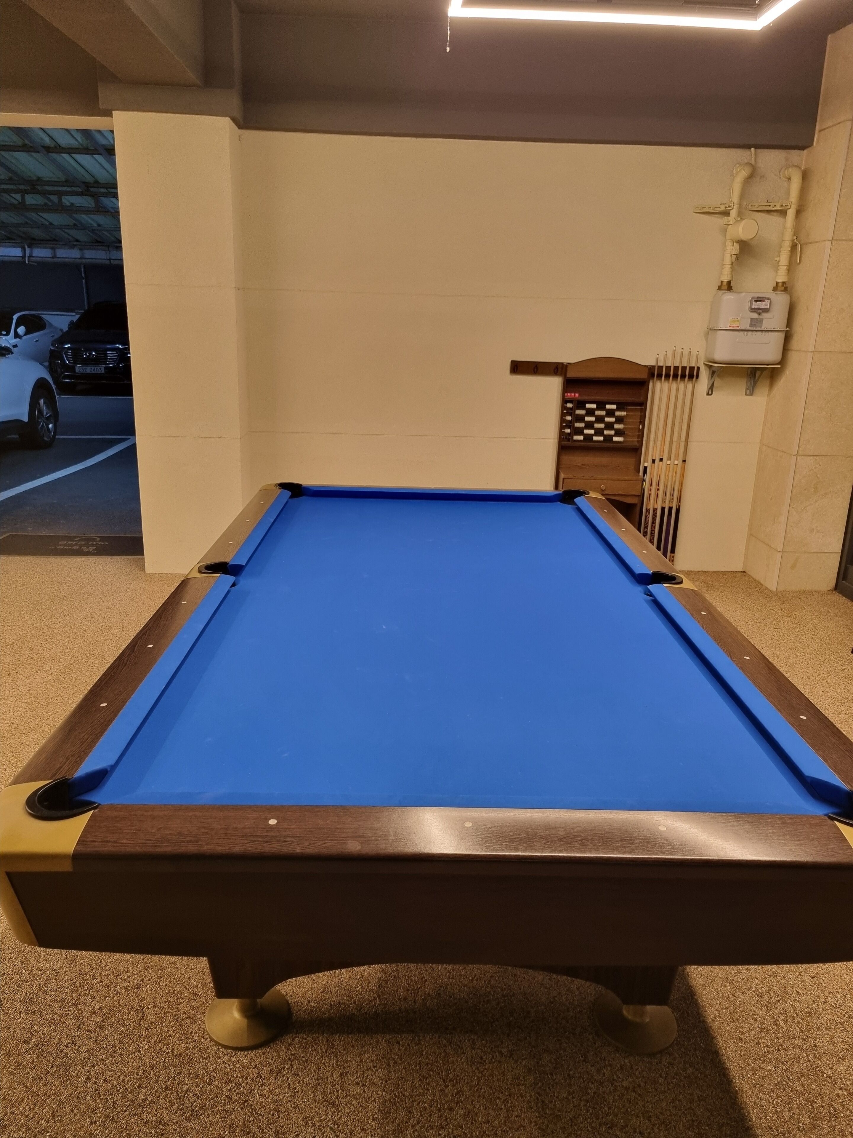 A pool table in a room with beige walls, a ceiling light, and a view of parked cars outside.