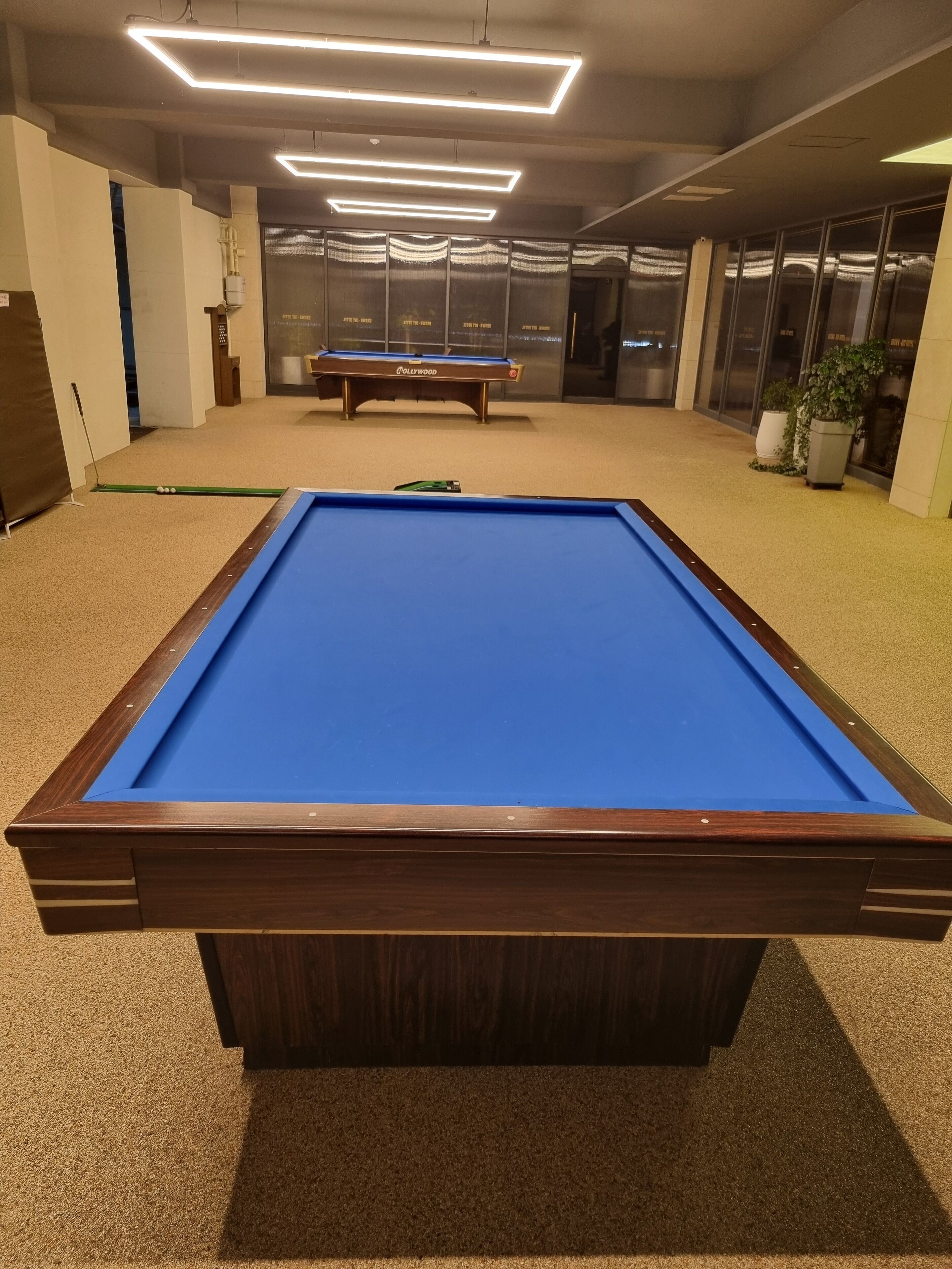 A pool table in a room with a carpeted floor and a ceiling with linear light fixtures.