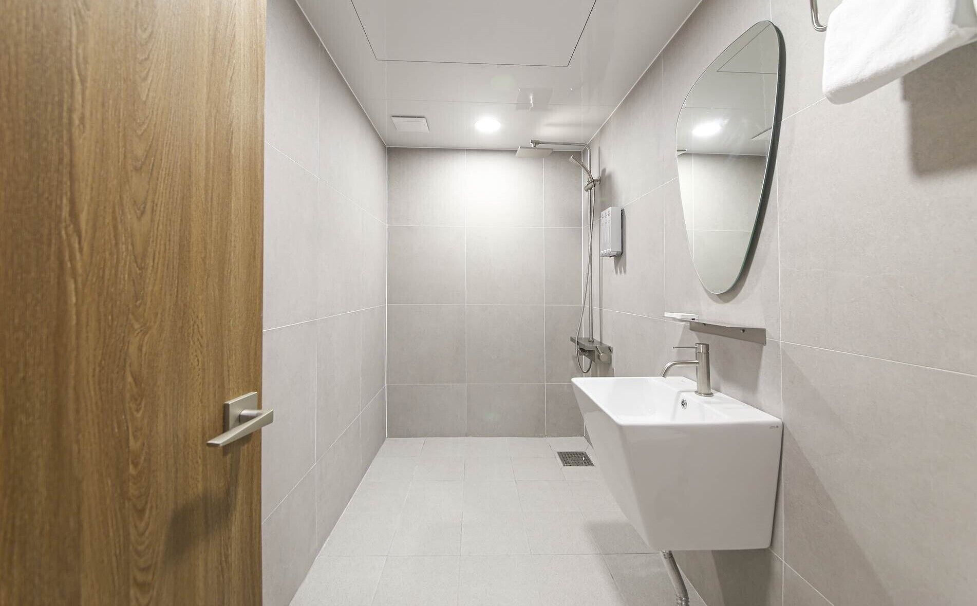 Un baño moderno con puerta de madera, lavabo blanco, espejo y zona de ducha.