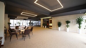 Un espacio interior amplio con piso alfombrado, varias mesas y sillas, y plantas en macetas.