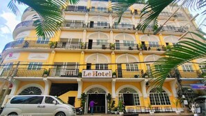 Un hôtel à plusieurs étages, doté de balcons, avec une enseigne indiquant « Le Hotel », et des voitures garées devant.