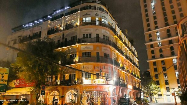 Un hôtel à plusieurs étages doté de balcons, une enseigne indiquant « Le Hotel », et une vue sur la rue avec d’autres bâtiments et des panneaux.