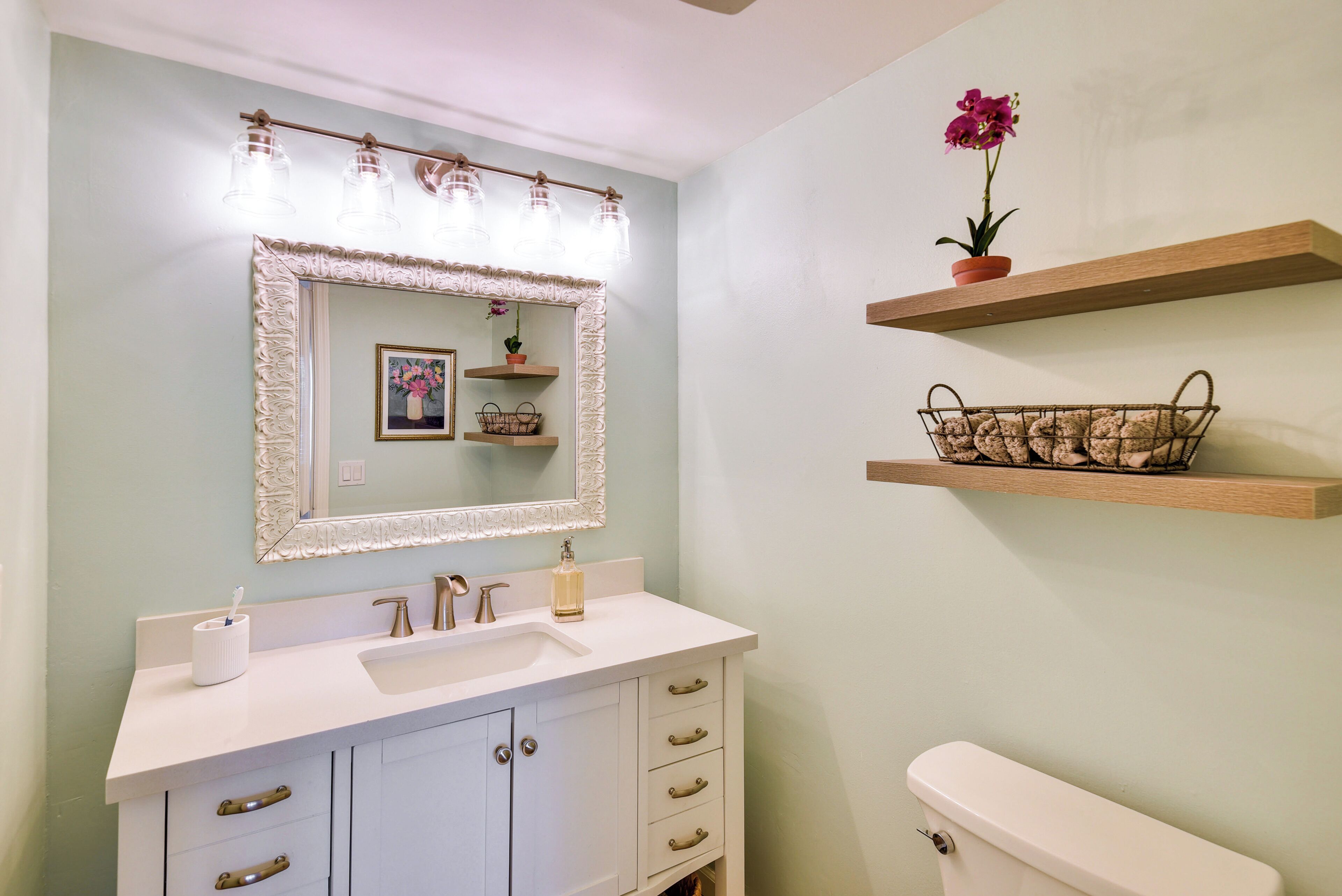 Un baño con un tocador blanco, un espejo grande y un inodoro. Hay repisas con objetos decorativos y una planta en maceta.