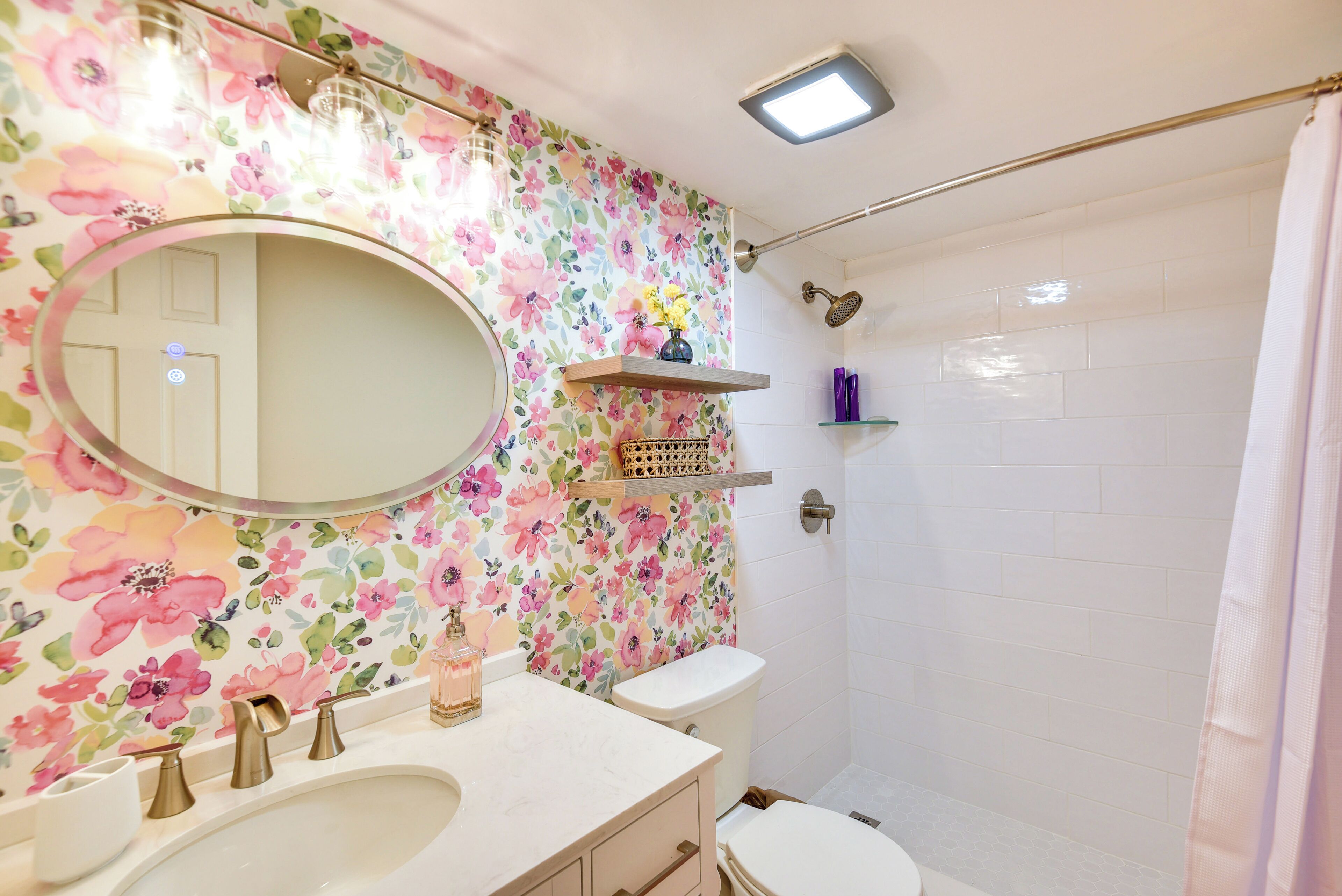 Une salle de bain avec un papier peint à motifs floraux, un miroir rond, un lavabo avec deux robinets, des toilettes et un espace douche.