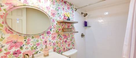 Un baño con papel tapiz floral, un espejo redondo, un lavabo con dos grifos, un inodoro y una zona de ducha.