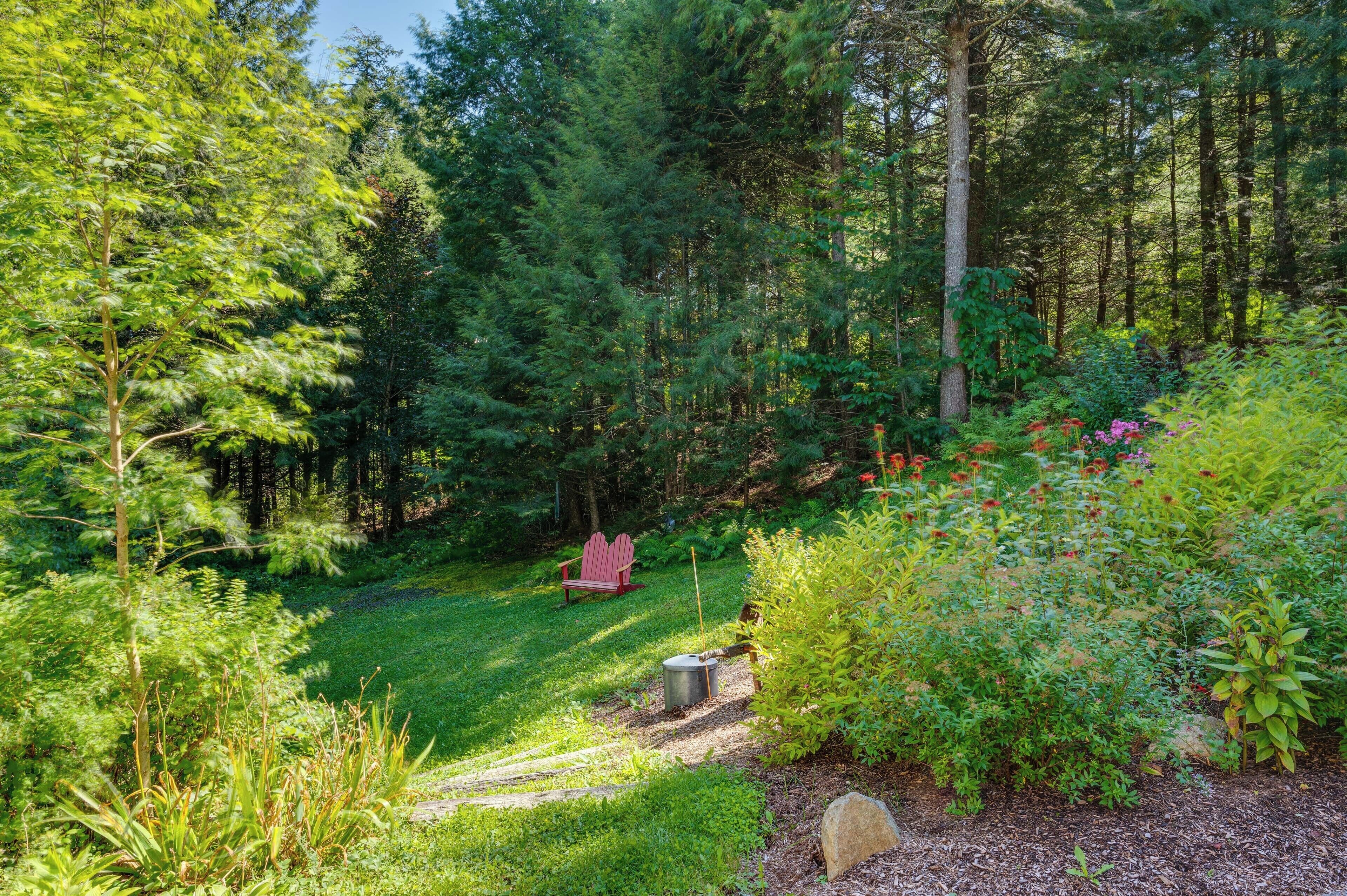 A forested garden with a red Adirondack chair, a metal trash can, and a stone.