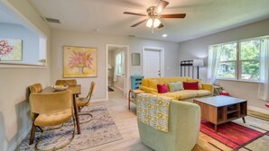 A living room with a sofa, coffee table, and chairs. There is a ceiling fan, a painting on the wall, and a door leading to another room.