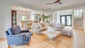 Ein geräumiges Wohnzimmer mit einer großen Couch, einem blauen Sessel und einem Couchtisch. Eine Deckenventilator, ein Essbereich mit einem Glastisch und eine Küche im Hintergrund sind ebenfalls zu sehen.