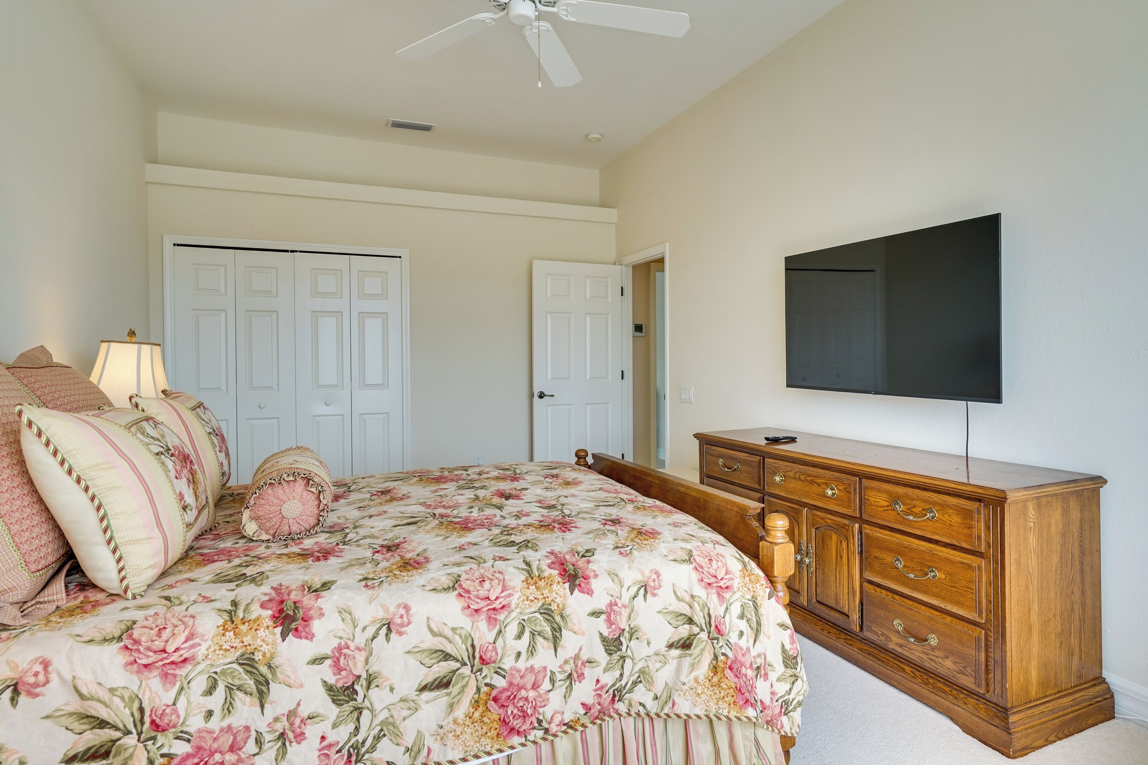 A bedroom with a floral bedspread, a wooden dresser, a flat-screen TV, and two white doors.