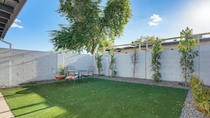 A backyard with artificial grass, a patio table and chairs, a small tree, and a white wall.