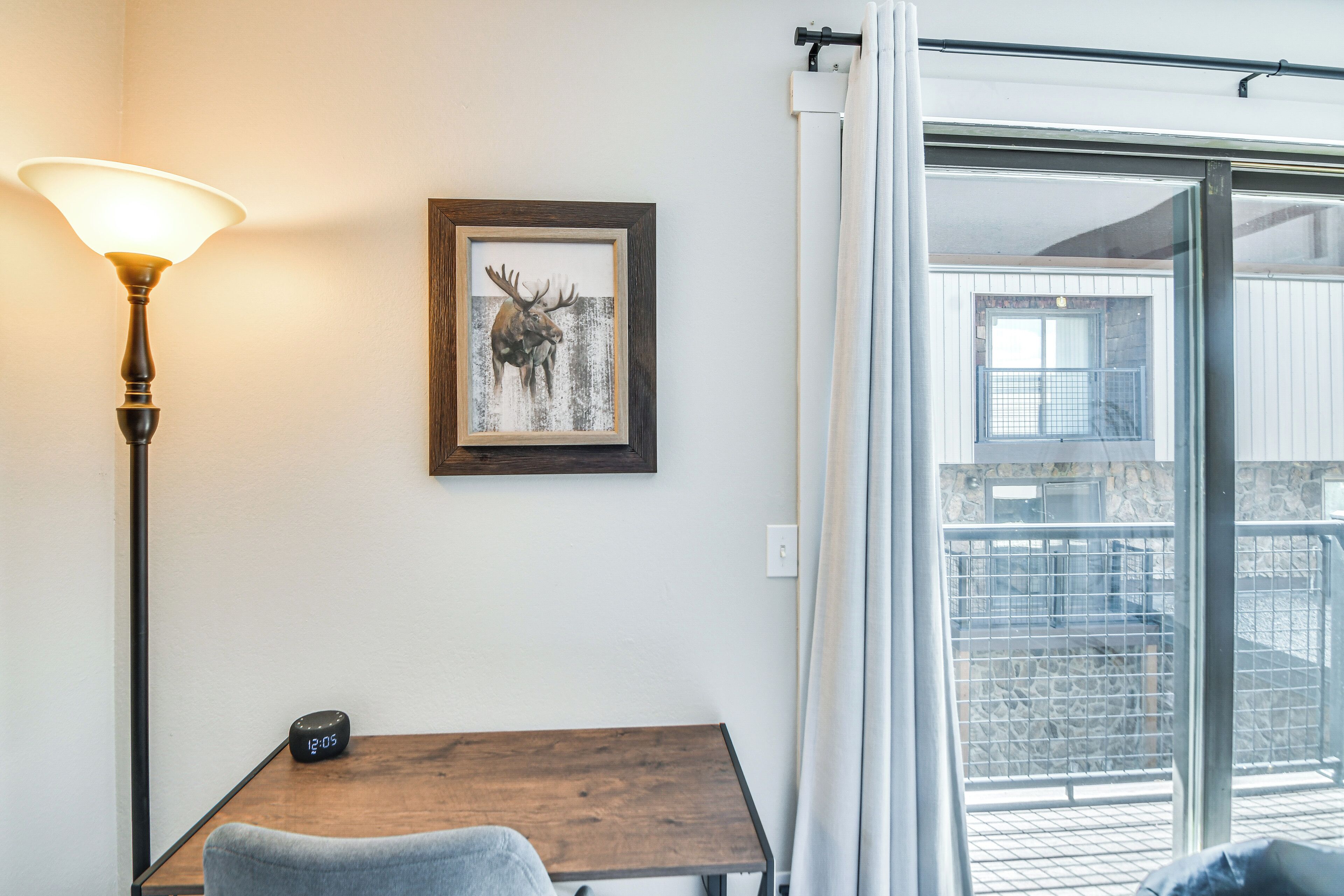 Una habitación con una mesa de madera, un cuadro enmarco de un alce, una lámpara y vistas a un espacio exterior por medio de una puerta de vidrio.