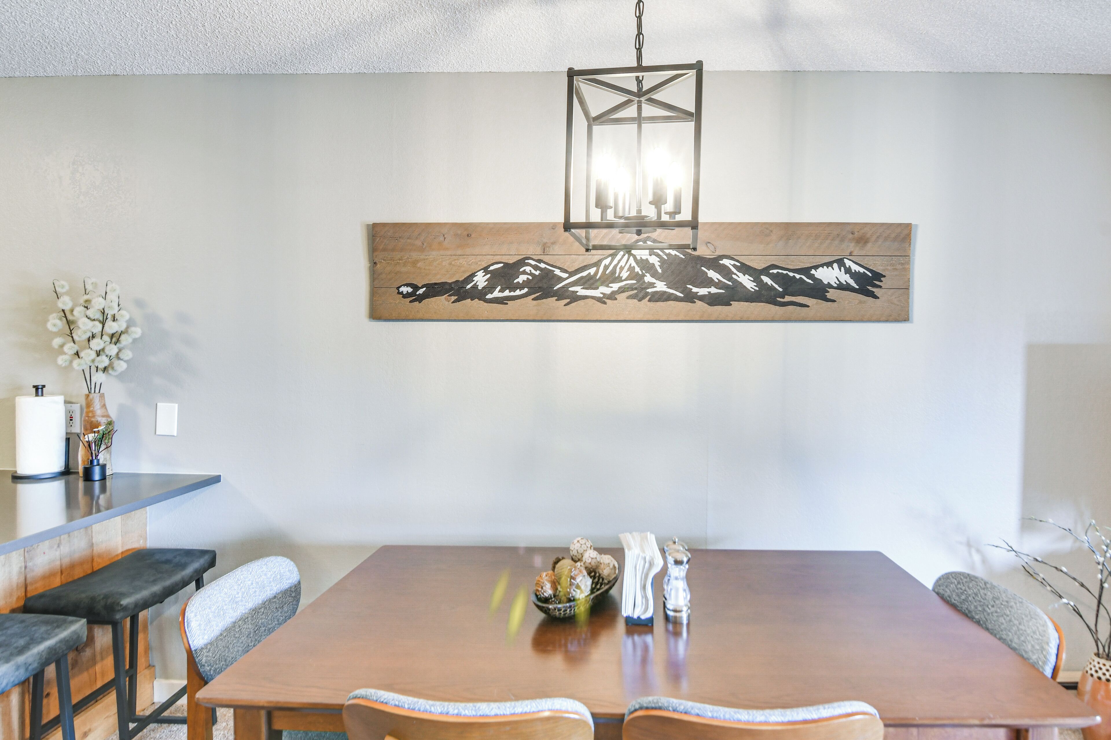 A dining area with a wooden table, grey upholstered chairs, a hanging lantern, and a wall-mounted artwork depicting mountains.
