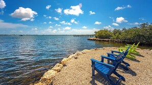 A beach with clear blue water, a wooden pier, and three blue chairs on a pebbled shore. - Captain House (Summerland Key)