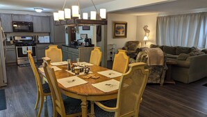 A dining area with a wooden table, chairs, and a chandelier. There is a kitchen in the background with modern appliances.