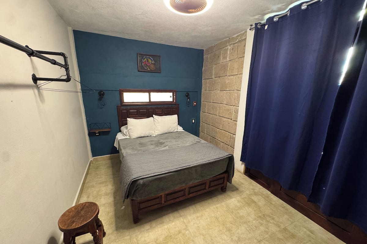 Ein Schlafzimmer mit Bett, Holz-Kopfteil, blauer Wand, Fenster, Vorhang und einem kleinen Hocker.