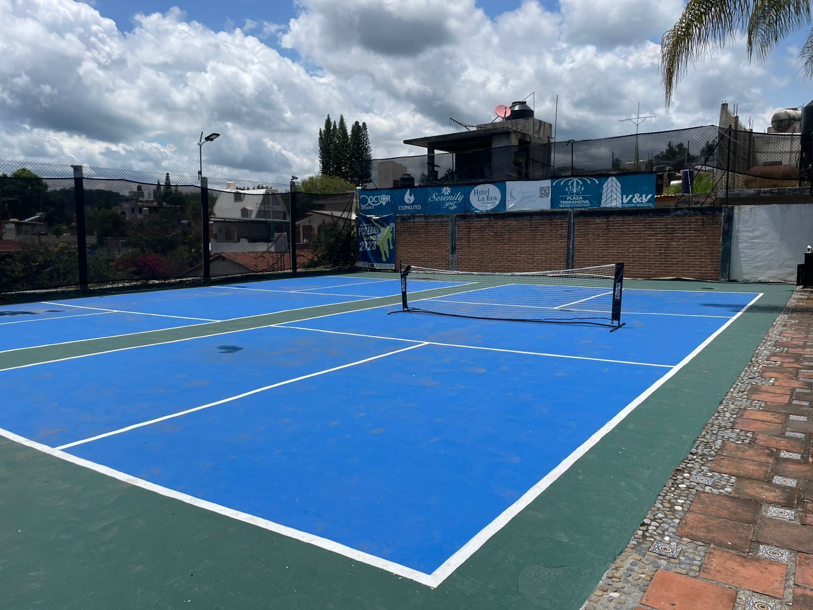 Una cancha de tenis exterior con red, rodeada por una cerca y anuncios de hoteles y negocios.