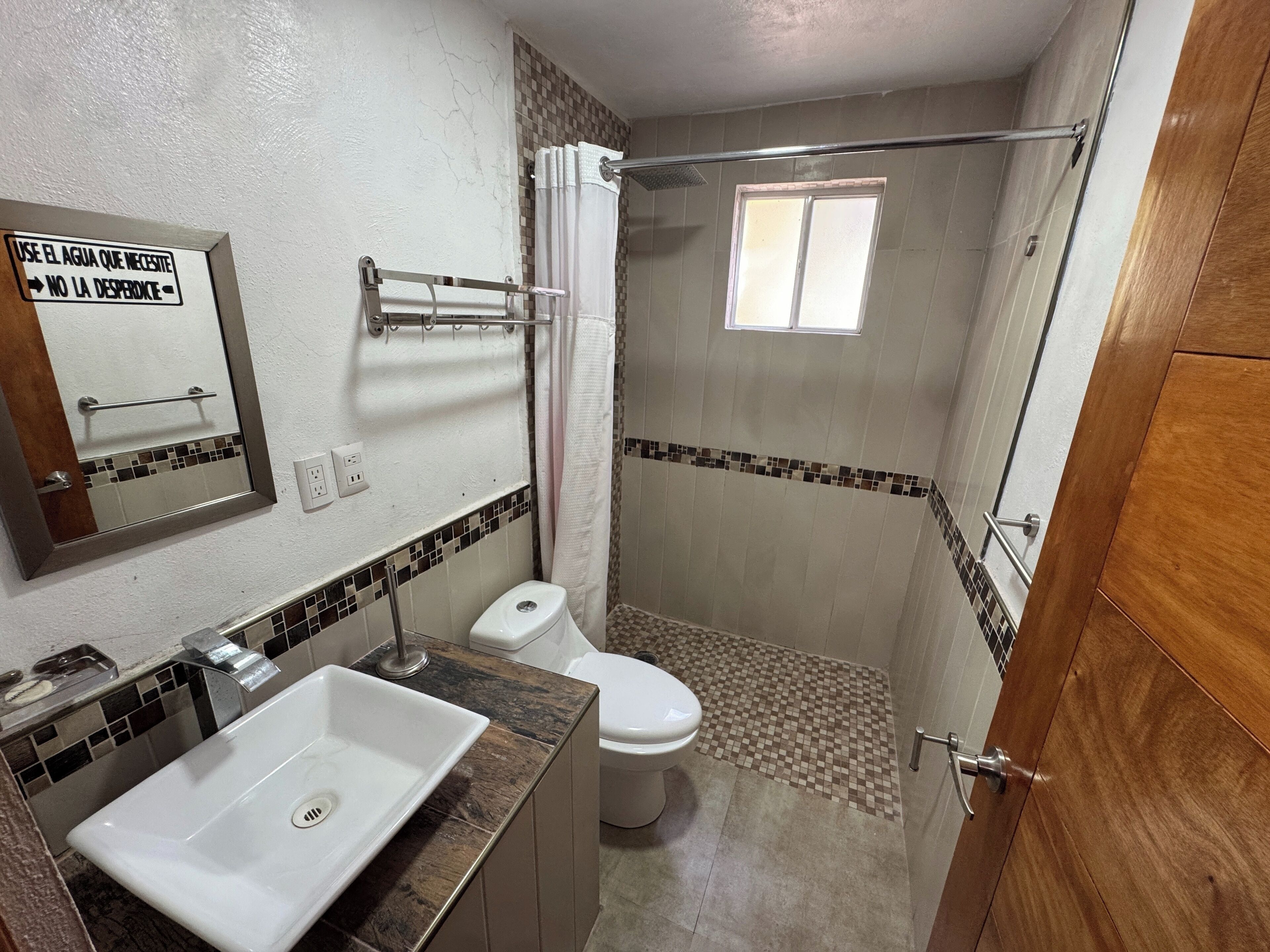 Un baño con lavamanos, inodoro y ducha blancos. Hay un espejo en la pared y una ventana sobre la ducha.