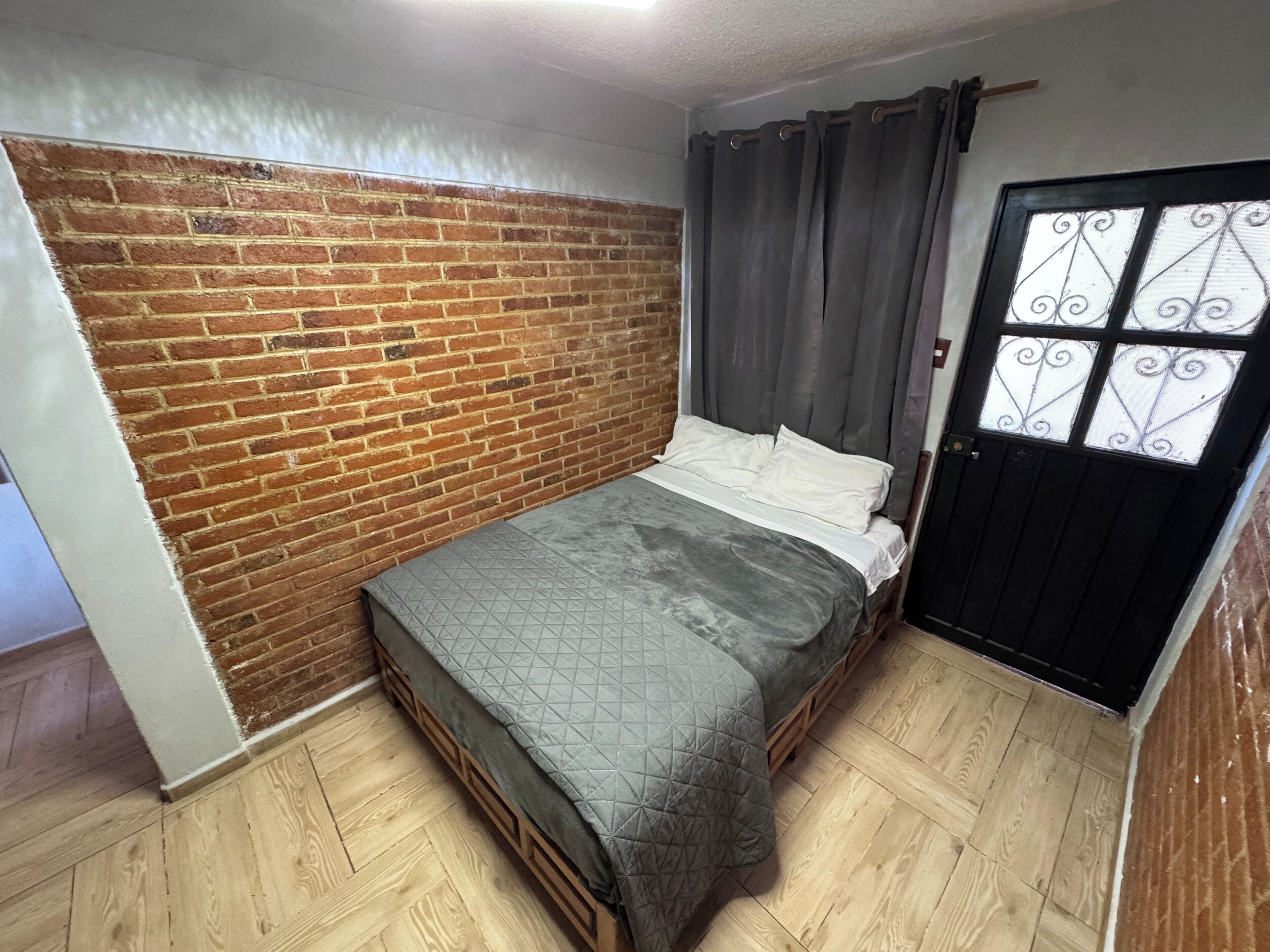 Een slaapkamer met een bakstenen muur, een bed met een grijze deken, houten vloeren en een zwarte deur met decoratieve glaspanelen.