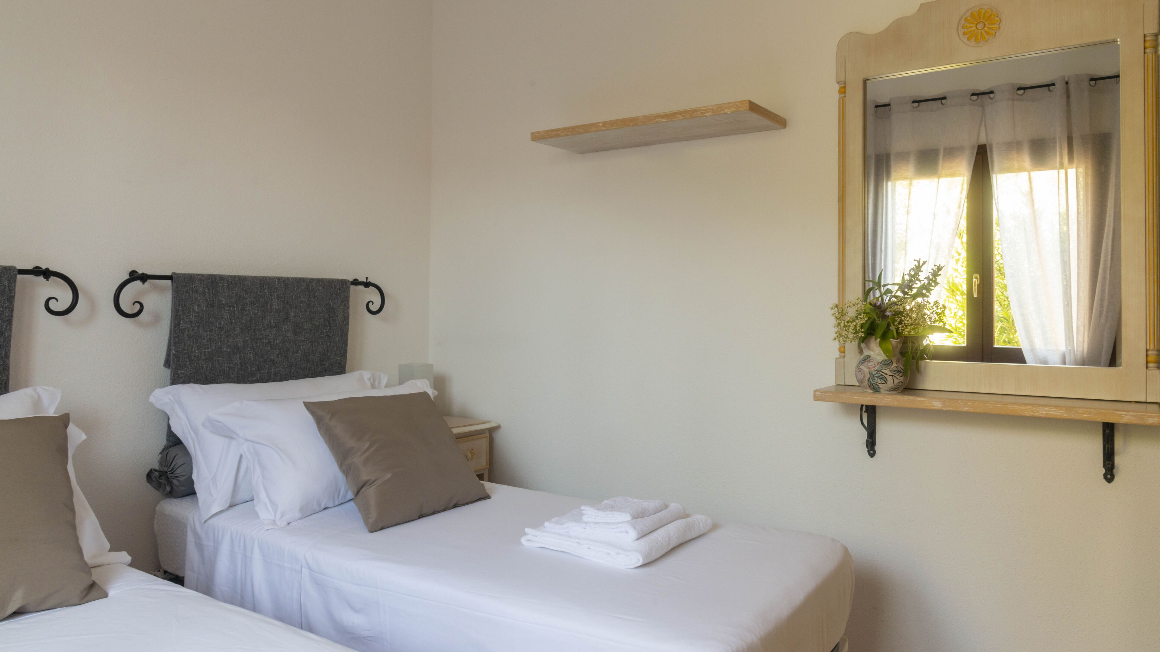 A hotel room with two beds, a small bedside table with a lamp, a mirror, and a window with curtains.