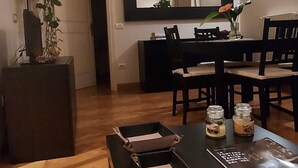 Een eetgedeelte met een zwarte tafel, stoelen en een houten vloer. Er staat een plant op een standaard, een televisie aan de muur en een airconditioning.
