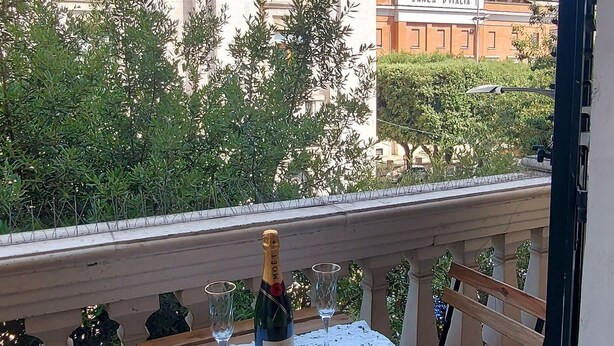 Een balkon met een tafel gedekt voor twee personen, een fles champagne en een glas. Op de achtergrond staat een gebouw met de tekst