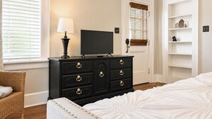 Une chambre à coucher comprenant un lit, une commode avec des tiroirs, un téléviseur, une lampe et une fenêtre avec des stores.
