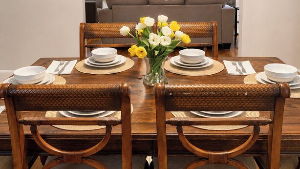 Un coin repas avec une table en bois dressée pour quatre personnes, entourée de chaises en osier, un vase avec des fleurs et un téléviseur en arrière-plan.