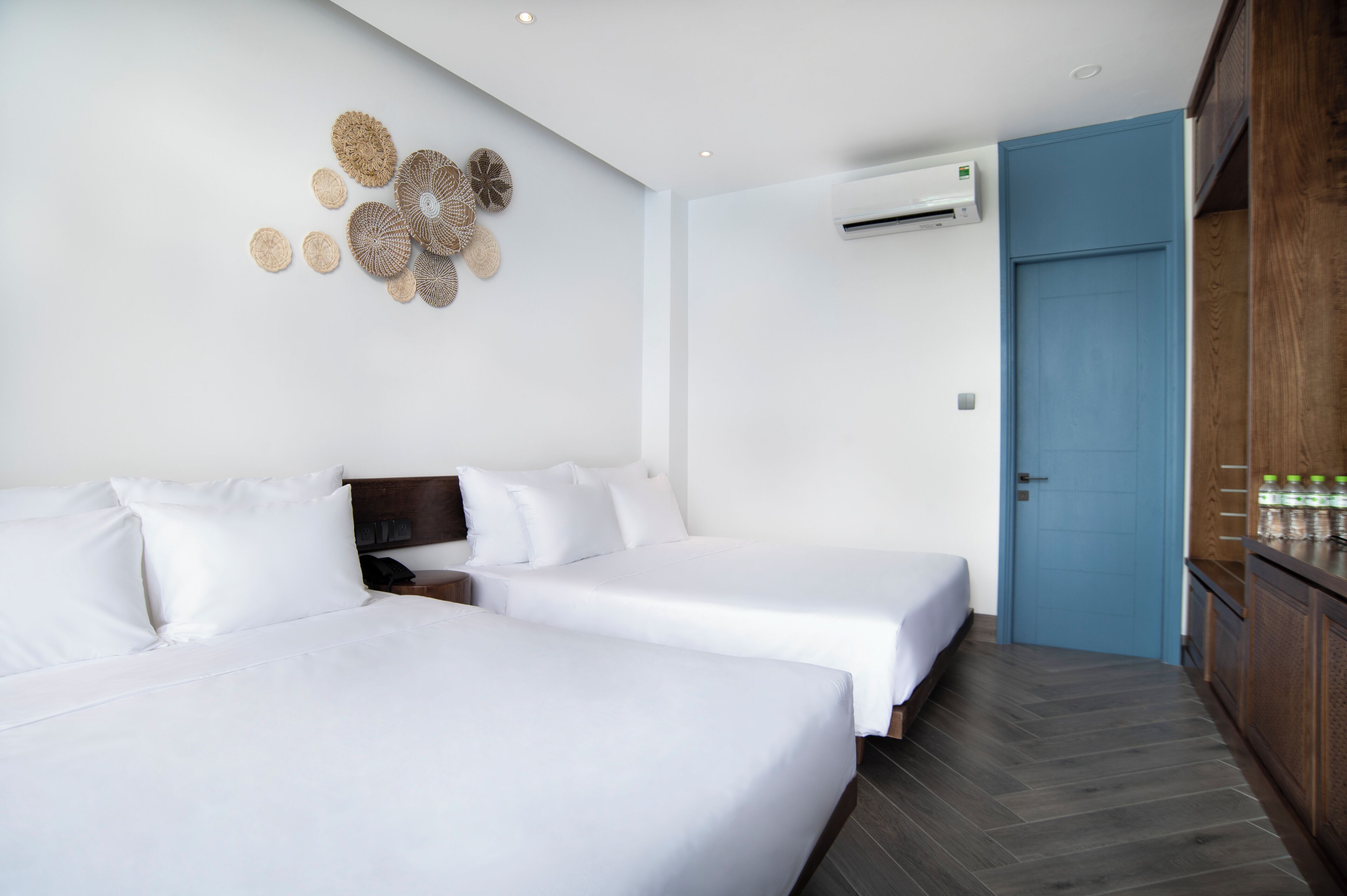 Ein Hotelzimmer mit zwei Betten, einer Wand-Klimaanlage, einer blauen Tür und einem Holzkoffer mit Flaschen.