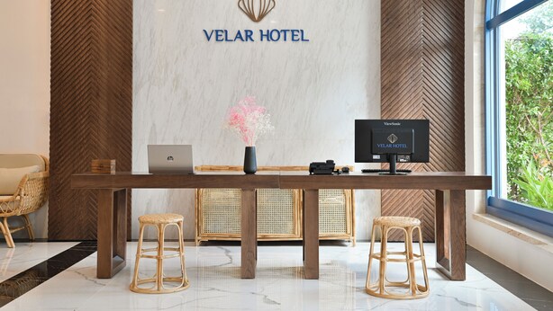 Un mostrador de recepción en VELAR HOTEL con una computadora portátil, un florero con flores rosadas y un monitor ViewStation.