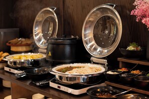 Un'area buffet con diversi piatti disposti in grandi pentole e ciotole, una cuociriso e un piccolo composizione floreale.