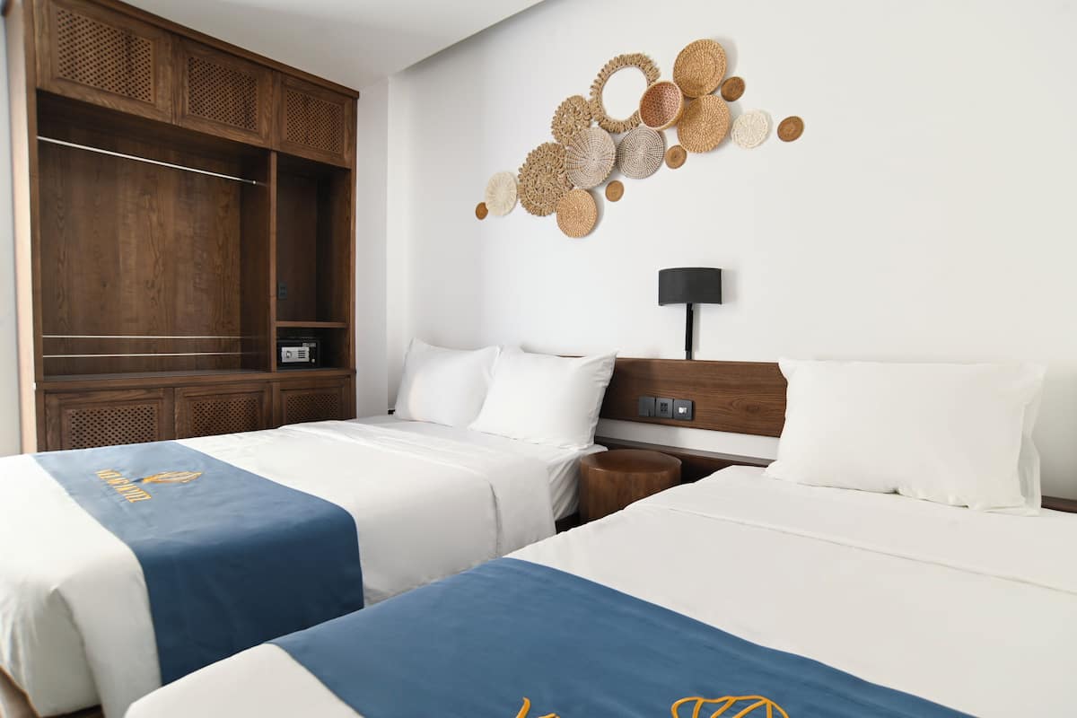 A hotel room with two beds, a wooden wardrobe, a wall-mounted lamp, and a decorative wall piece.